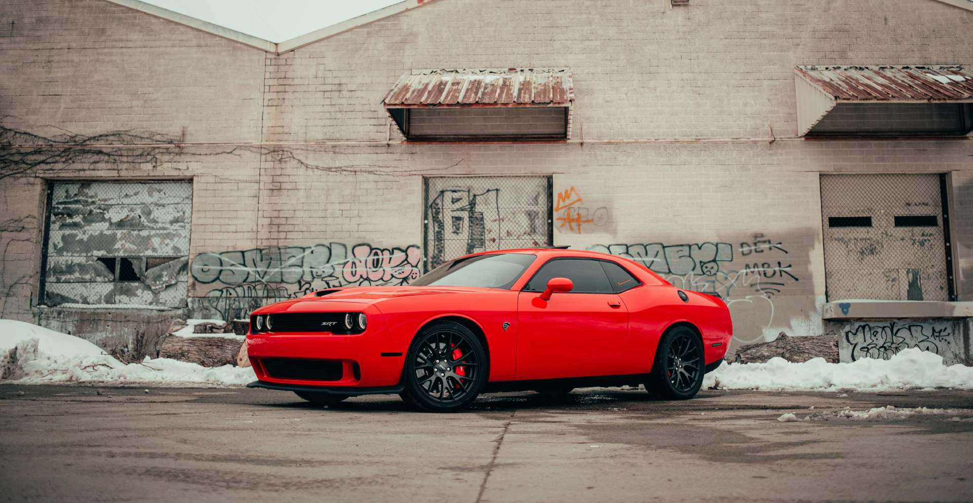 A vibrant red car is parked in front of a weathered brick building covered in graffiti