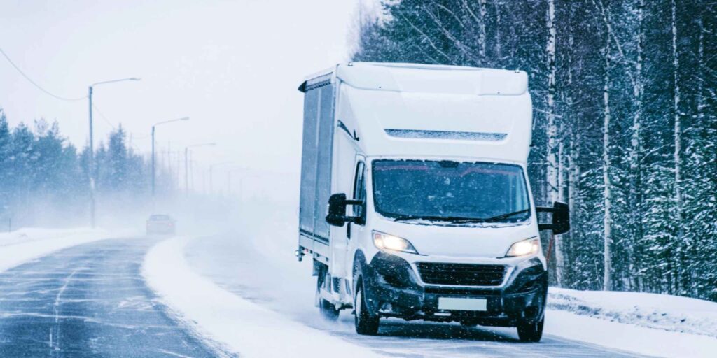 A white delivery truck drives cautiously on a snowy, winding road during a blizzard, with its headlights on
