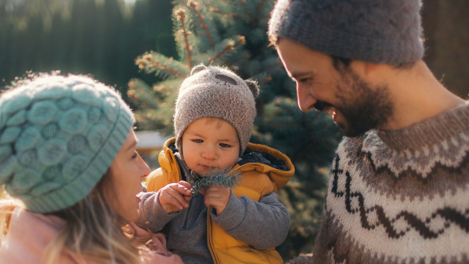 A young family wearing knit hats and sweaters stands near a pine tree outdoors, with the toddler in a yellow jacket curiously holding a pine needle sprig