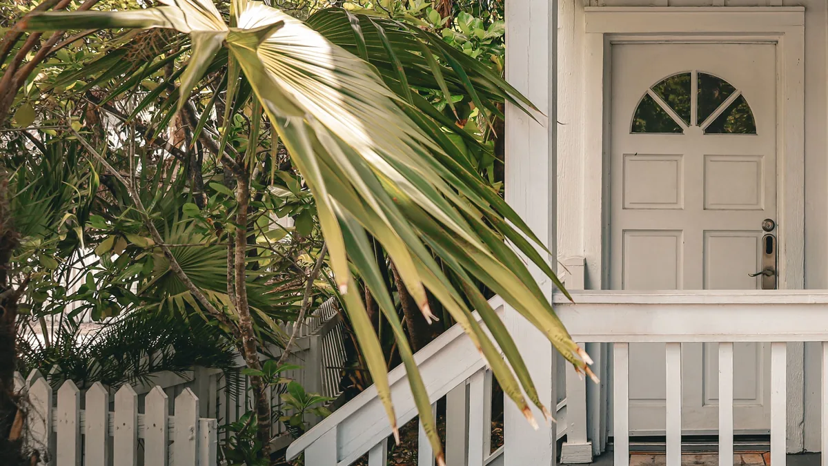 A white wood frame cottage porch with an arched window in the front door, white railings, a picket fence and palm fronds